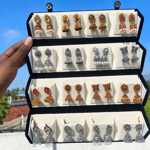 Hand holding an open black velvet box with 16 pairs of antique gold and oxidised silver jhumka earrings – Jaan Creations