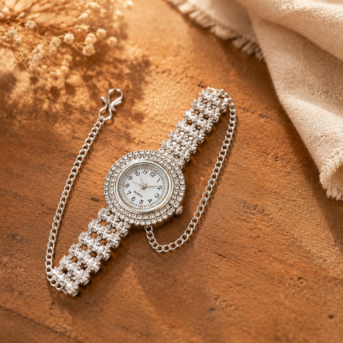 A silver-tone watch with a diamond-encrusted bezel and matching link bracelet, styled on a warm wooden surface with dried flowers and a beige fabric corner.