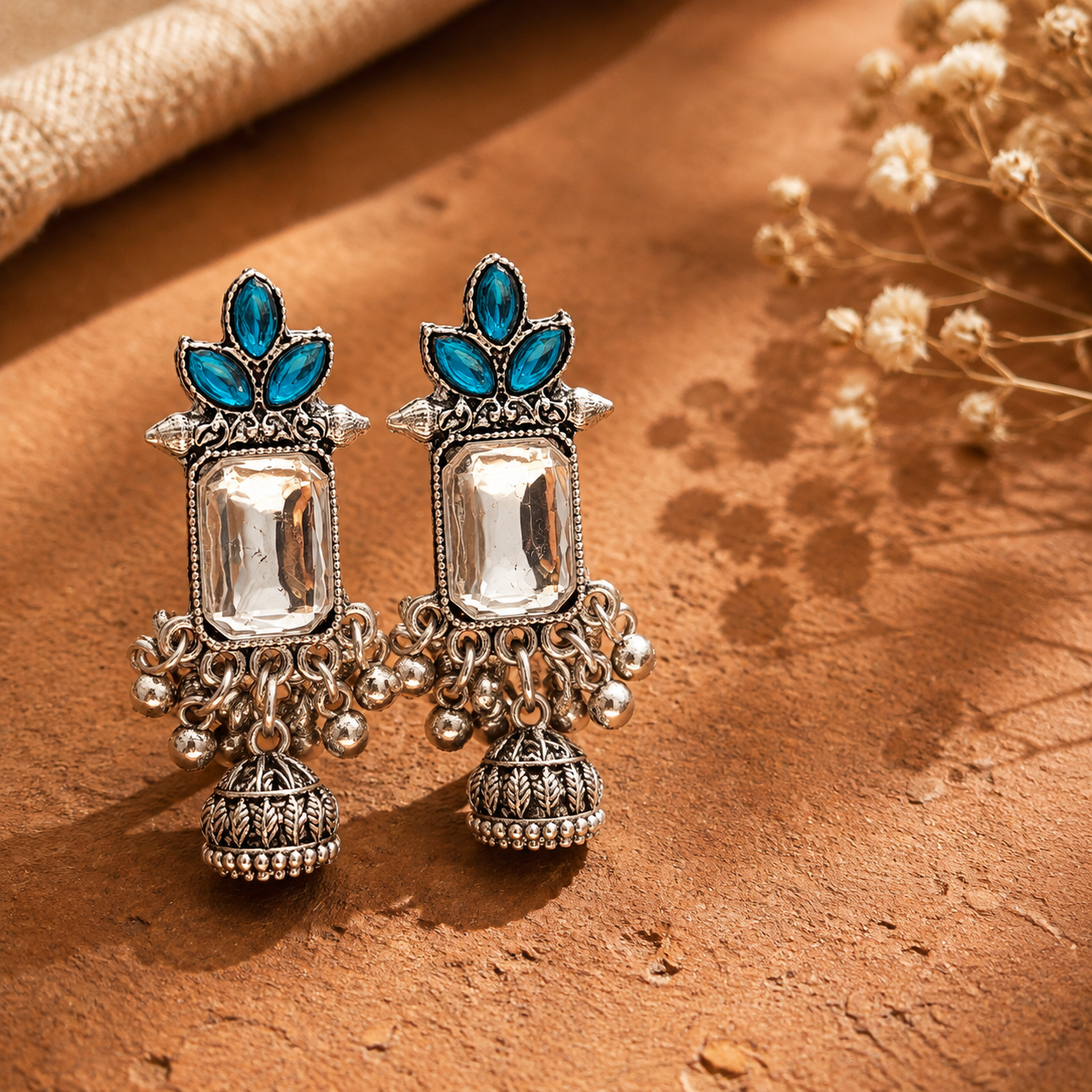 A pair of ornate silver-toned Jhumka earrings with turquoise blue stones and clear rectangular center stones, displayed on a textured brown surface with dried flowers in the background.