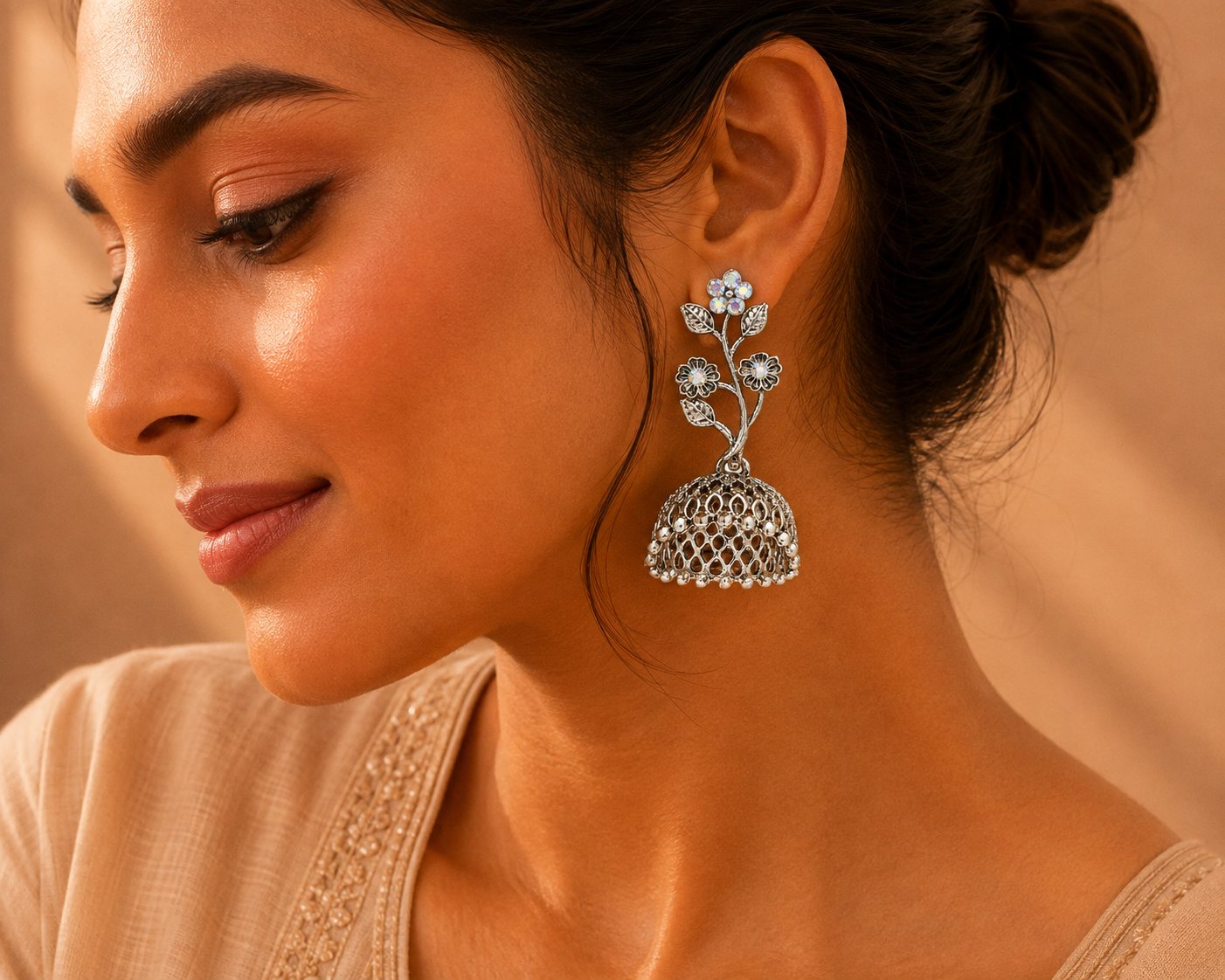 A close-up side profile of a woman wearing a silver-toned jhumka earring with floral motifs and clear stones, set against a warm beige background.