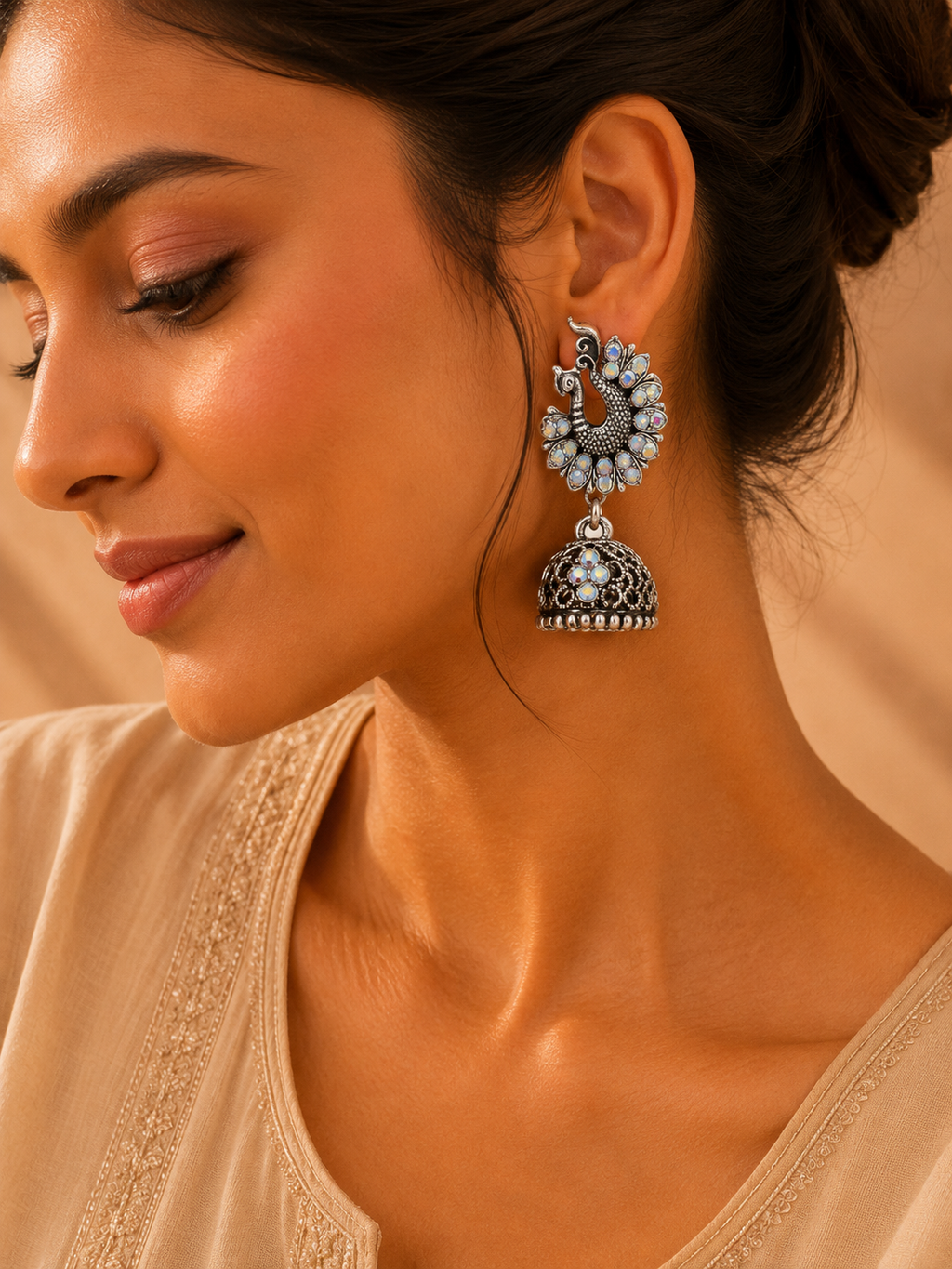 A close-up of a woman wearing a pair of ornate silver-toned jhumka earrings with light blue stone accents, set against a warm beige background.