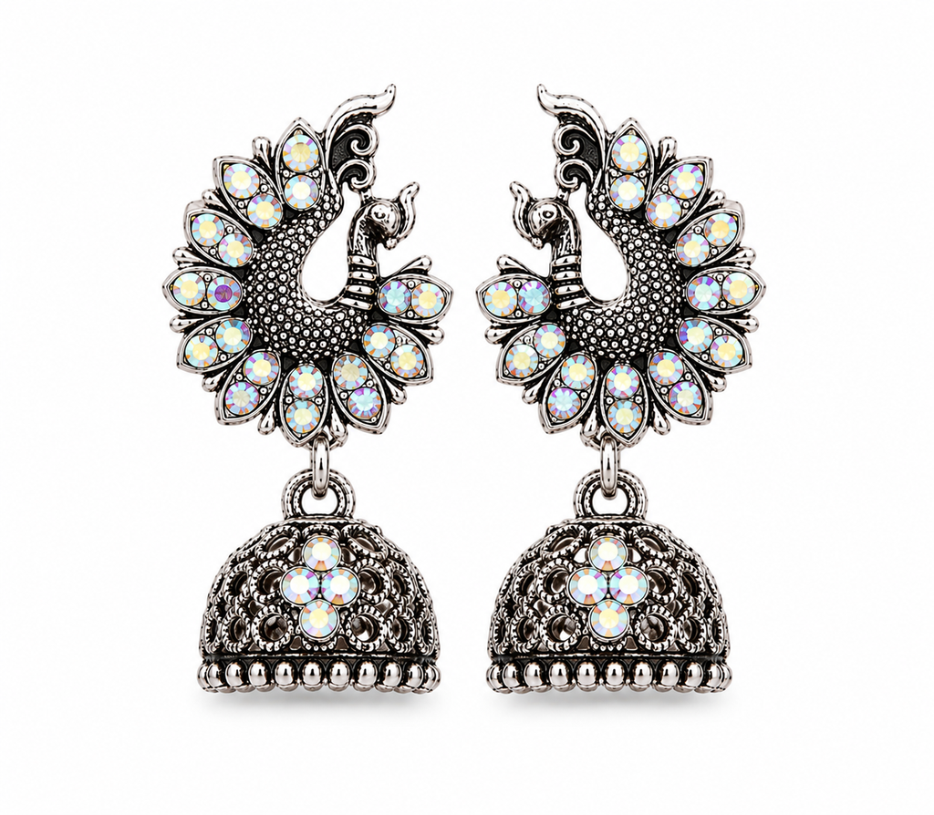 A pair of silver-toned Jhumka earrings featuring peacock motifs adorned with iridescent white and multicolor stones, hanging from ornate filigree bell-shaped drops on a plain white background.