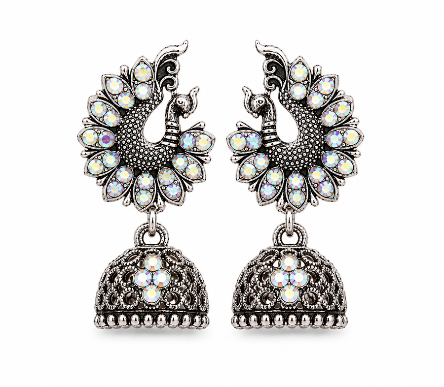A pair of silver-toned Jhumka earrings featuring peacock motifs adorned with iridescent white and multicolor stones, hanging from ornate filigree bell-shaped drops on a plain white background.