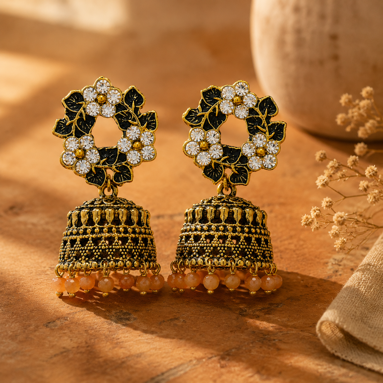 A pair of ornate Indian-style jhumka earrings with gold-plated bell-shaped drops, black enamel floral motifs, clear rhinestones, and peach-colored bead fringe, displayed on a warm wooden surface.
